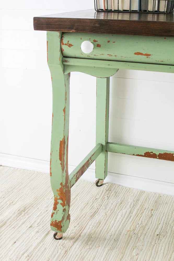 Vintage table with green distressed paint and wooden top on a white floor.