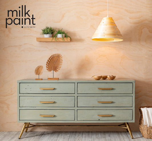 Green dresser with gold accents against a wooden wall with 'milk paint' branding.