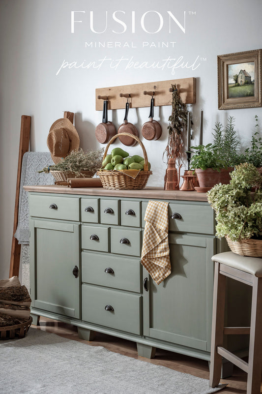 Green kitchen cabinet with Fusion Mineral Paint branding, featuring pots, a basket, and plants.