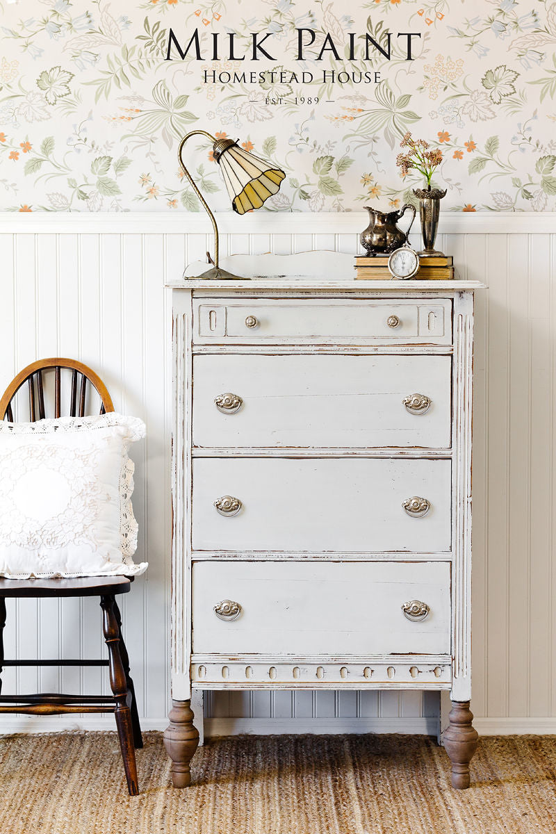White distressed dresser with decorative items against a floral wallpapered wall, featuring 'Milk Paint Homestead House' branding.