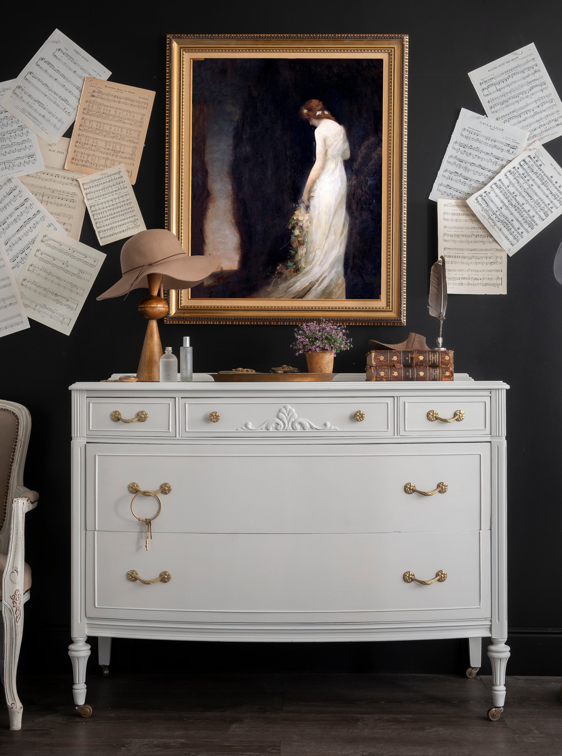 White dresser with gold accents against a dark wall with framed artwork and papers.