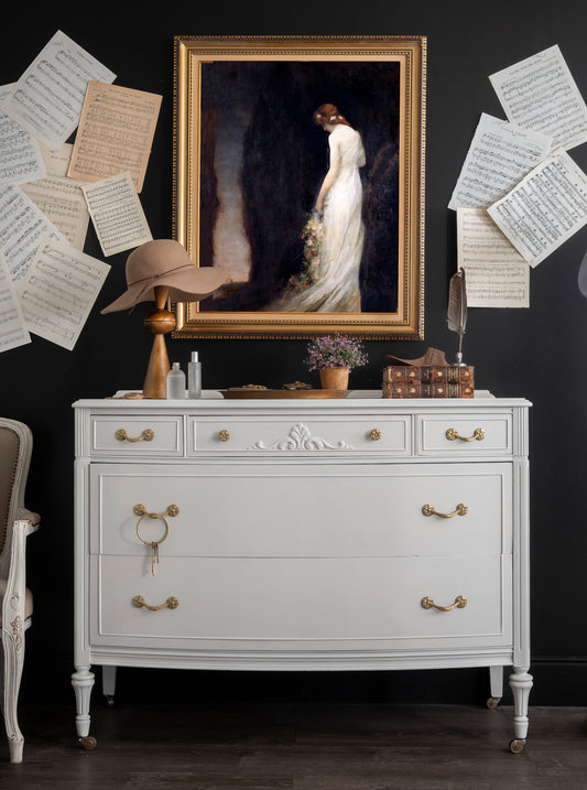 White dresser with gold accents against a dark wall with framed artwork and papers.