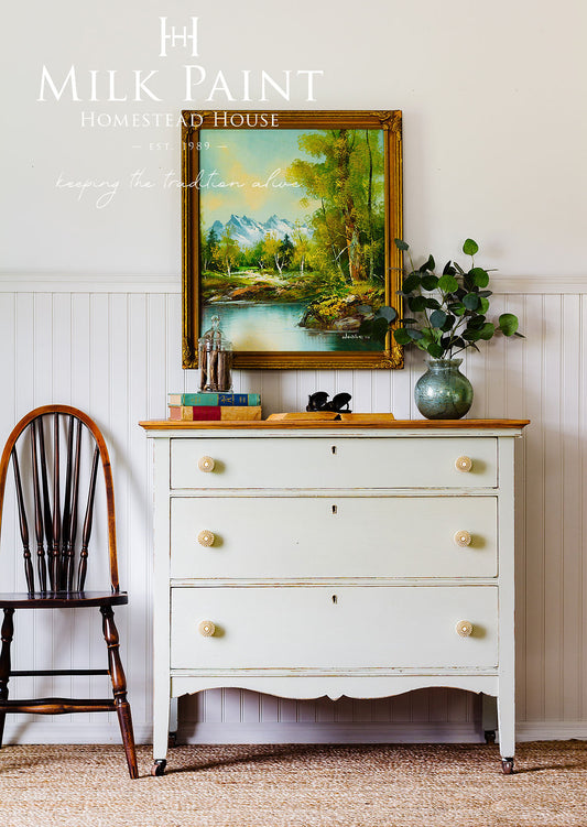 White dresser with gold accents, framed painting, and decorative items against a white wall.
